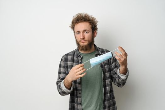 Getting rid protective medical mask young handsome man holding it stretching it in front of face ready wearing plaid shirt and olive t-shirt under. White background.