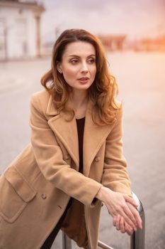 Beautiful Parisian woman standing outdoors posing leaned on handrail for camera in an autumn beige coat, beautiful hair with urban city background. Vertical portrait. Warm tinted photo.