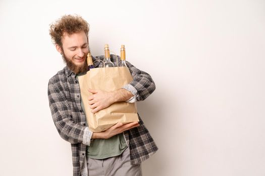 Young man carrying paper shopping bag with bottles of wine smiling looking down on it. Handsome young man with curly hair in olive t-shirt looking at camera isolated on white background.