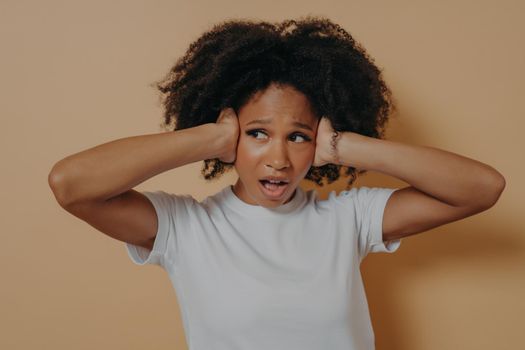 Young annoyed african woman covering her ears and keeping mouth opened, feeling stressed, does not want to hear you while standing isolated on beige background, unhappy female suffering from headache
