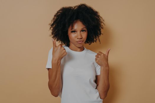 Success sign. Confident young african woman showing thumbs up with both hands and looking at camera isolated on beige background, showing winner gesture and smiling. Body language concept
