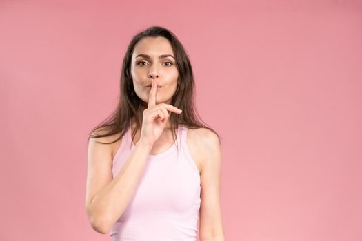 Gesture with a finger to the mouth says to be quiet or keep a secret young woman with dark hair in trendy pink t-shirt looking at camera. Facial expressions, emotions, feelings.