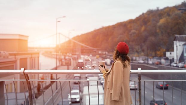 Young woman warming up with cup of coffee standing backwards on a pedestrian bridge looking away. Portrait of stylish young woman wearing autumn coat and red beret outdoors. Toned image.