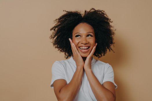 Joyful young dark skinned woman feeling happy, smiling at camera and touching cheeks with both hands, carefree african female in white tshirt expressing positivity, posing isolated on beige background