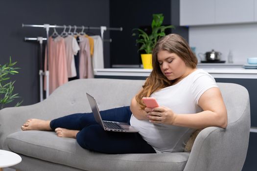 Texting or checking on social media while working on laptop sitting at home curve body young woman staying at home during quarantine. Self isolation. Working distantly from home using laptop.