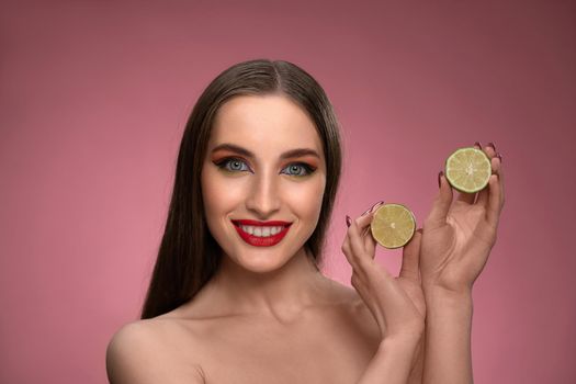 Happy young woman show lime benefit to health, holding two parts sliced in her hands looking charming. Charming joyful funny lady with long hair isolated on pink background.