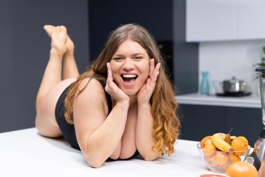 Overweight girl with big breasts laying on the kitchen table. An obese young sexy chubby white girl in black swimsuit at modern kitchen with fruits next to her.