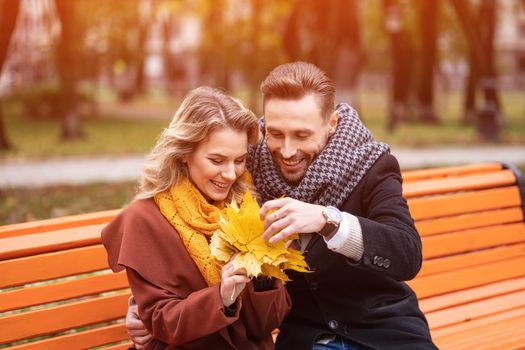 Young embracing happy romantic couple sitting gently hugged on a bench in park wearing coats and scarfs. A romantic couple sitting on a bench hugging in the park. Love story.