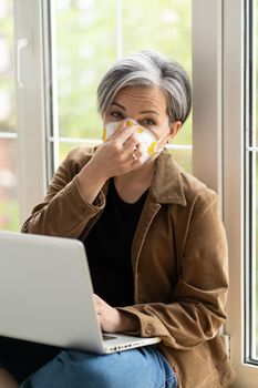 Mature woman in protective fp1 face mask working on laptop compute sitting on a windowsill at free woking zone or workstation. A beautiful aged woman runs her own business as a freelancer.