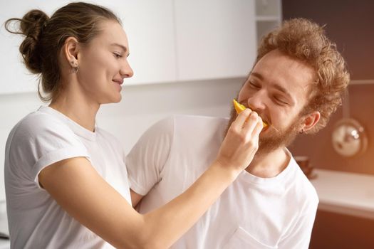 Pretty girl feeds or nursing her boyfriend. Beautiful young couple feeding each other, making fun at modern kitchen and smiling while cooking at home.