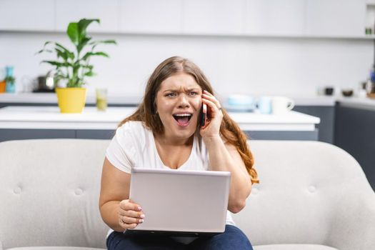 Mad screaming talking on the phone young fat girl working on laptop sitting on the sofa at home with the kitchen on background. Self acceptance. Working distantly from home using laptop.