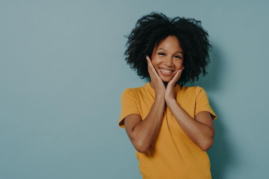 Smiling cute mixed race young woman in yellow tshirt touching cheeks with hands and feeling happy after hearing compliment from boyfriend, standing isolated on blue background with copy space