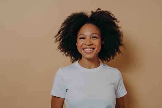 Beautiful young african female smiling cheerfully at camera and demonstrating happiness, standing against color wall in studio,dark skinned woman in casual outfit with curly hair showing white teeth