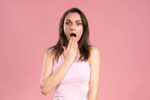 Astonished young woman looking surprised with dark hair in pink t-shirt looking at camera with holding hand at the lips. Facial expressions, emotions, feelings.