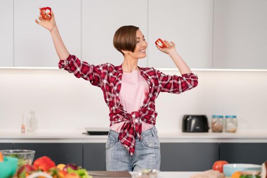 Pretty woman dancing holding cut bell pepper in hands in red shirt playing with food, while cooking in the white kitchen. Healthy food at home. Healthy food leaving concept. New house concept. 