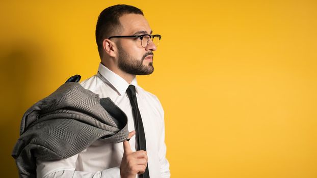 Handsome serious business man with trendy beard dressed in greyish suite posing holding his jacket on his shoulder hanging it behind looking isolated on yellow background.