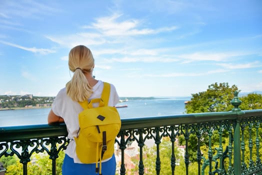 Blond girl with a backpack looking at the river Saint Lawrence at Quebec Canada Sanny day by Chateau Frontenac Castle