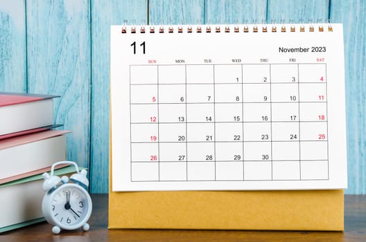 November 2023 Monthly desk calendar for the organizer to plan 2023 year with alarm clock and books on wooden table.