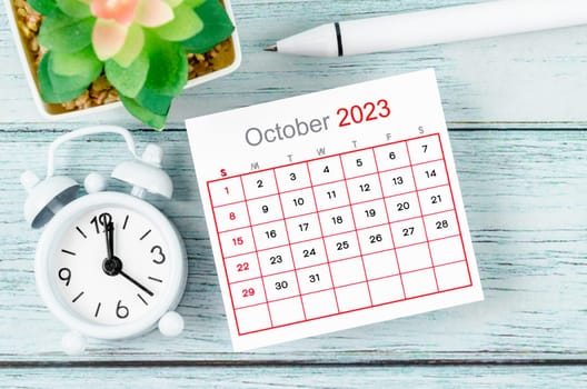 October 2023 Monthly calendar year and alarm clock with pen on blue wooden background.