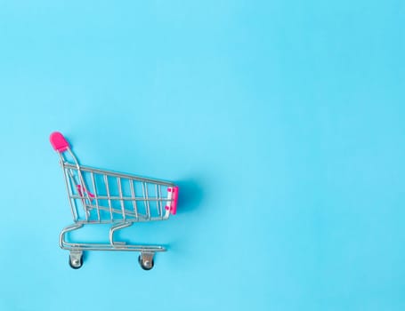 Empty toy metal supermarket shopping cart on blue background with copy space.