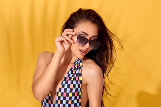 Portrait of young pretty tanned tattooed woman with long brunette hair, wearing checkered swimsuit with neckline and dark sunglasses posing on yellow background. She touches her sunglasses