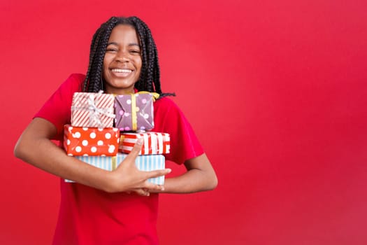 Happy african woman holding lots of colorful gifts in studio with red background