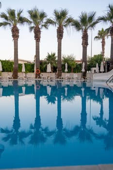 Luxurious hotel with swimming pool, palm trees, sun loungers and seating area. Palm trees are reflected in the clear water of the pool.