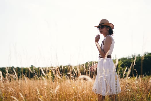 Portrait of young woman walking among high grasses in summer meadow wearing straw hat and linen dress enjoying nature. Harmony and balance.