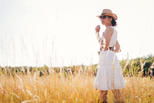 Portrait of young woman walking among high grasses in summer meadow wearing straw hat and linen dress enjoying nature. Harmony and balance.