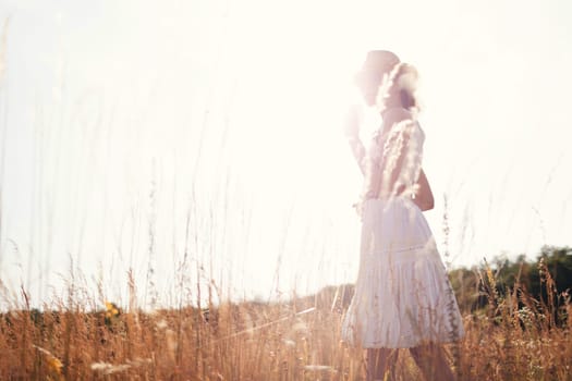 Portrait of young woman walking among high grasses in summer meadow wearing straw hat and linen dress enjoying nature. Harmony and balance.