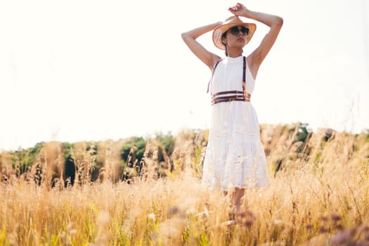 Spirit of freedom. An attractive boho girl in blouse, hat and sunglasses standing on the field on the background of a blue sky. Summer vacation, traveling. Bohemian, modern hippie style