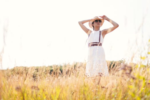 Spirit of freedom. An attractive boho girl in blouse, hat and sunglasses standing on the field on the background of a blue sky. Summer vacation, traveling. Bohemian, modern hippie style