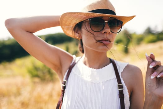 Spirit of freedom. An attractive boho girl in blouse, hat and sunglasses standing on the field on the background of a blue sky. Summer vacation, traveling. Bohemian, modern hippie style