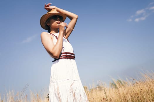 Spirit of freedom. An attractive boho girl in blouse, hat and sunglasses standing on the field on the background of a blue sky. Summer vacation, traveling. Bohemian, modern hippie style