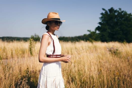 Spirit of freedom. An attractive boho girl in blouse, hat and sunglasses standing on the field on the background of a blue sky. Summer vacation, traveling. Bohemian, modern hippie style