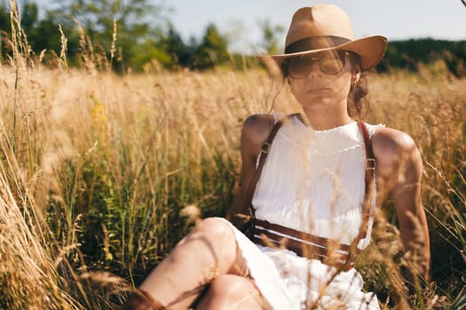 Spirit of freedom. An attractive boho girl in blouse, hat and sunglasses standing on the field on the background of a blue sky. Summer vacation, traveling. Bohemian, modern hippie style