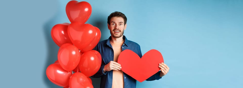 Valentines day concept. Man falling in love, looking startled at girlfriend, showing big red heart and standing near balloon over blue background.