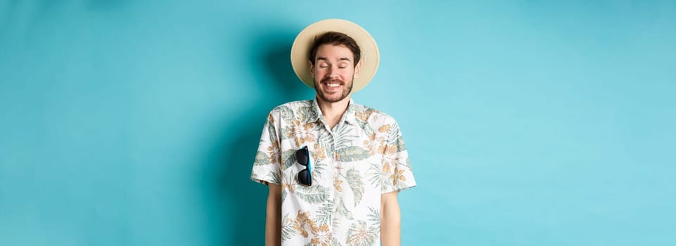 Happy tourist enjoying summer holiday, wearing straw hat and hawaiian shirt, standing joyful on blue background.