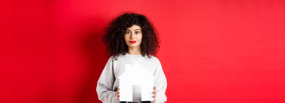 Real estate. Young caucasian woman in casual clothes showing paper house cutout, buying property or renting apartment, standing on red background.