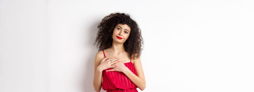 Touched young woman with curly hair, wearing red dress, holding hands on heart and smiling grateful, say thank you, standing over white background.