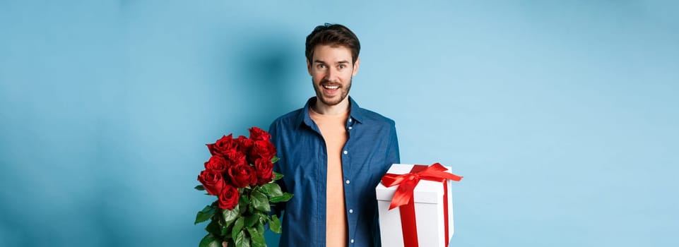 Love and relationship concept. Happy young man bring flowers and gift on romantic date. Boyfriend wish bouquet of roses and present standing on blue background.