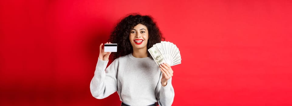 Portrait of stylish young woman with curly hair, showing money in cash and plastic credit card, red background.