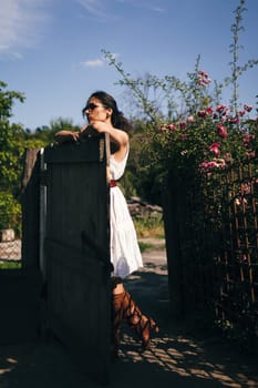 Young fashionable woman stands near fence in the garden or backyard of countryside house. Summer time, routine, relax, harvest concept.