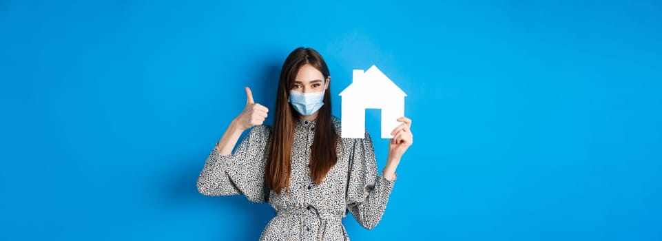 Real estate, covid-19 and pandemic concept. Candid woman in medical mask showing thumb up and paper house cutout, reanting apartment, standing on blue background.