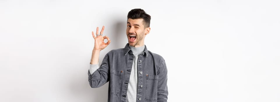 Excited handsome guy with moustache, winking and showing okay sign, smiling pleased, assure all good, praise nice work, making good job gesture, standing against white background.