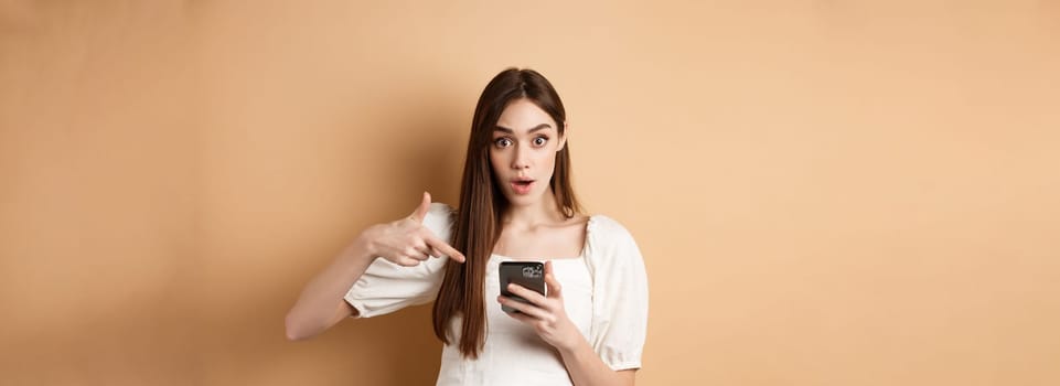 Online shopping concept. Excited girl pointing at smartphone screen and looking at camera amazed, standing against beige background.