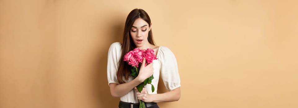 Valentines day. Surprised girl receive gift from lover on date, looking amazed at beautiful bouquet of flowers, holding roses on beige background.