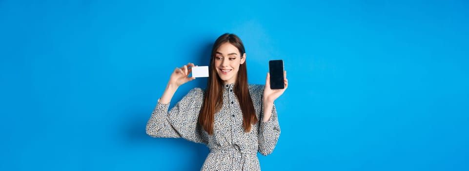 Online shopping. Beautiful female model showing empty cellphone screen and plastic credit card, smiling pleased, buying in internet store, blue background.
