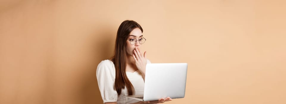 Shocked girl look at laptop screen, covering mouth with amazed face, standing in glasses on beige background.
