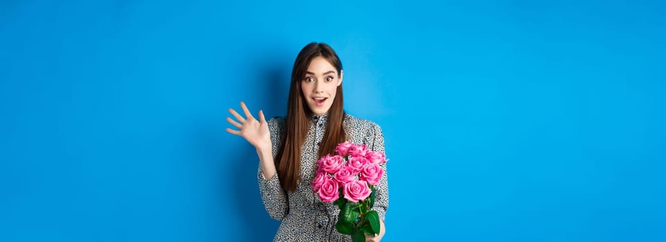 Valentines day concept. Surprised pretty girl holding bouquet of flowers from lover, looking amazed and happy at camera, standing on blue background.
